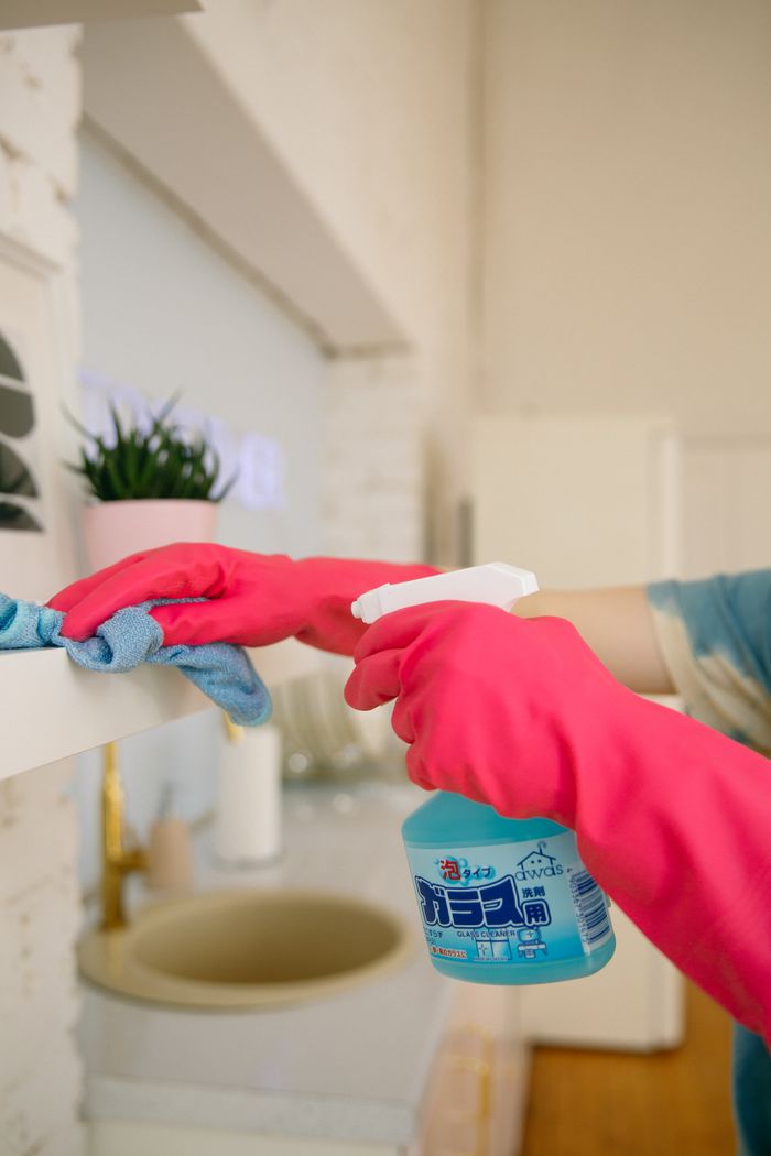 about-us-01 Person using spray bottle and cloth for cleaning a kitchen shelf, ensuring hygiene and cleanliness.